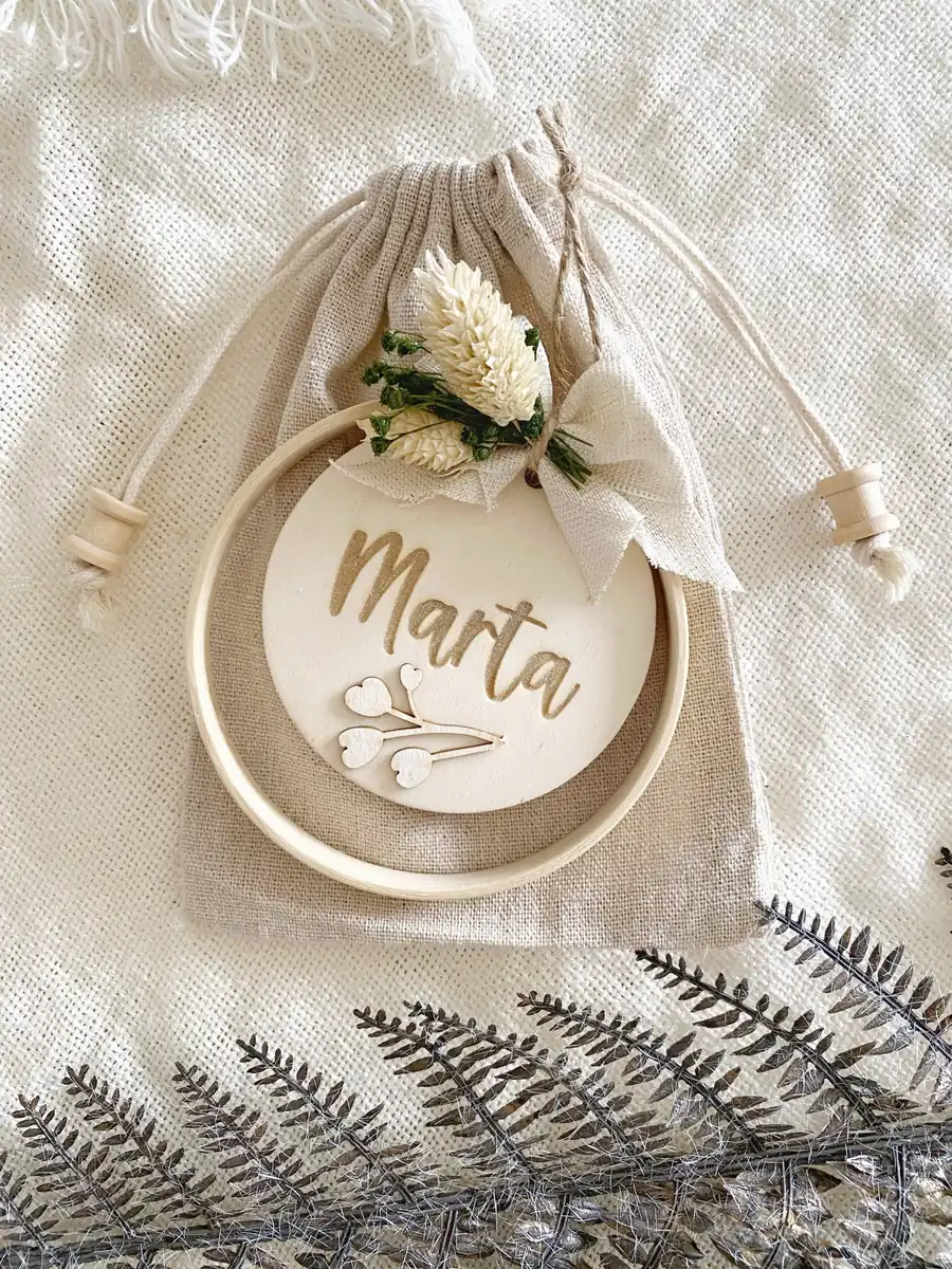 Bastidor decorativo personalizado con nombre del invitado, lazo de lino y flores secas sobre bolsa de lino natural con tapones de madera