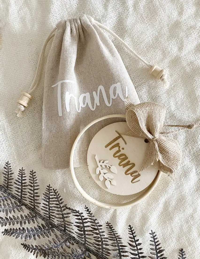 Bastidor decorativo con nombre y motivo de hojas en madera junto a bolsa de lino con cordón y tapones de madera