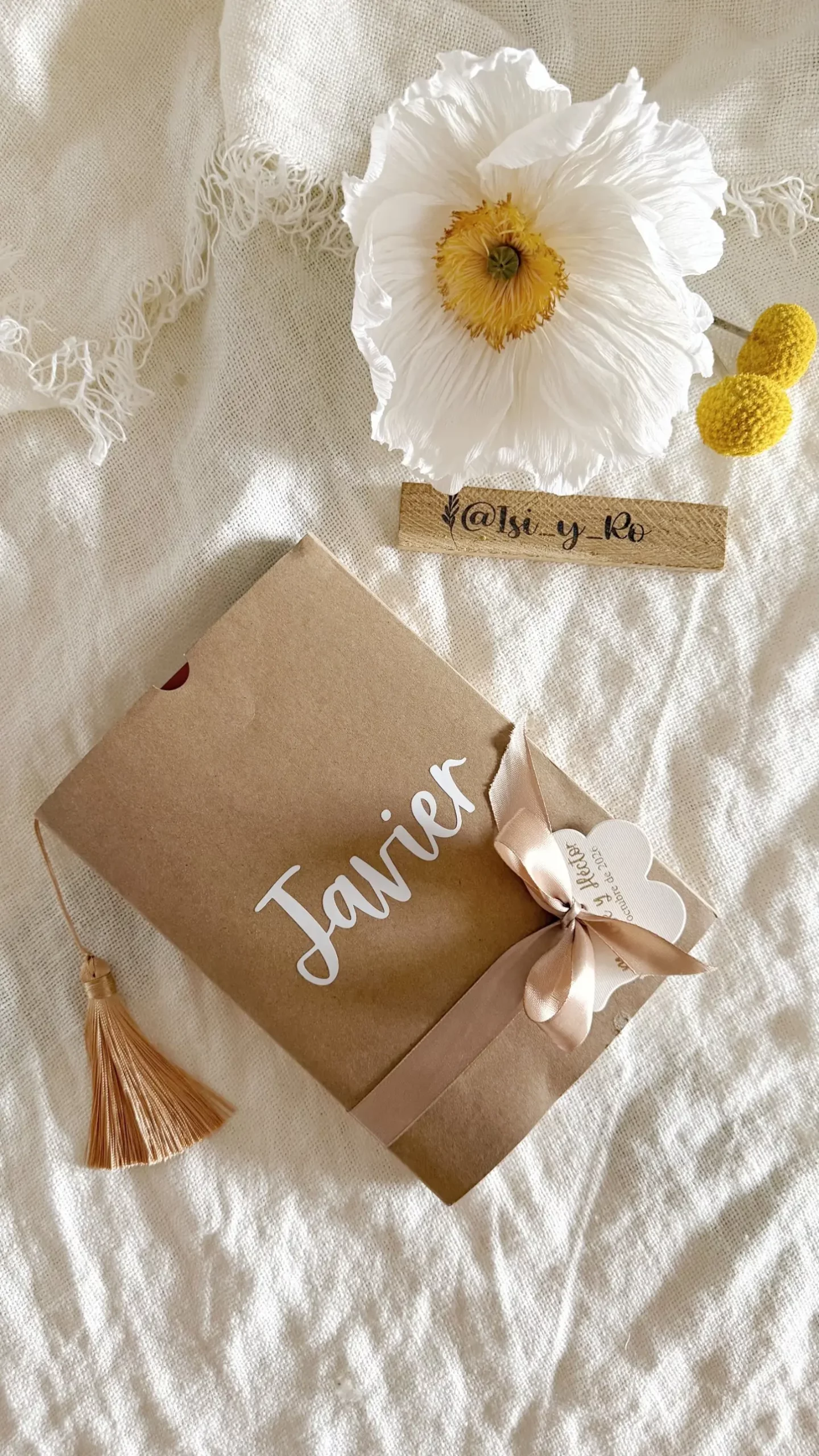 Funda kraft con nombre en vinilo blanco, lazo de raso, etiqueta en forma de flor y borla color camel